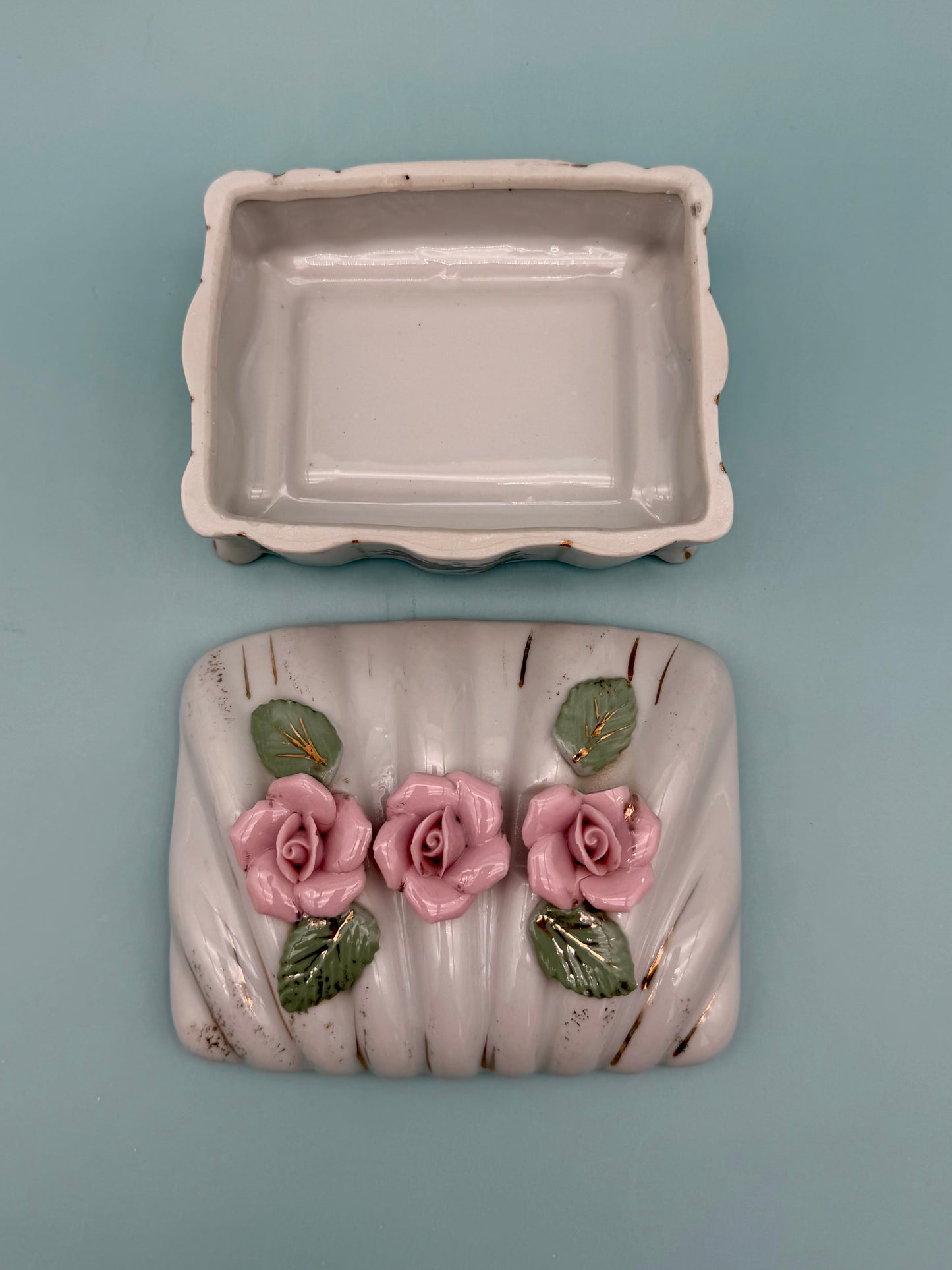 Open porcelain stash box with empty ivory interior beside its rose-topped lid, teal and pink background