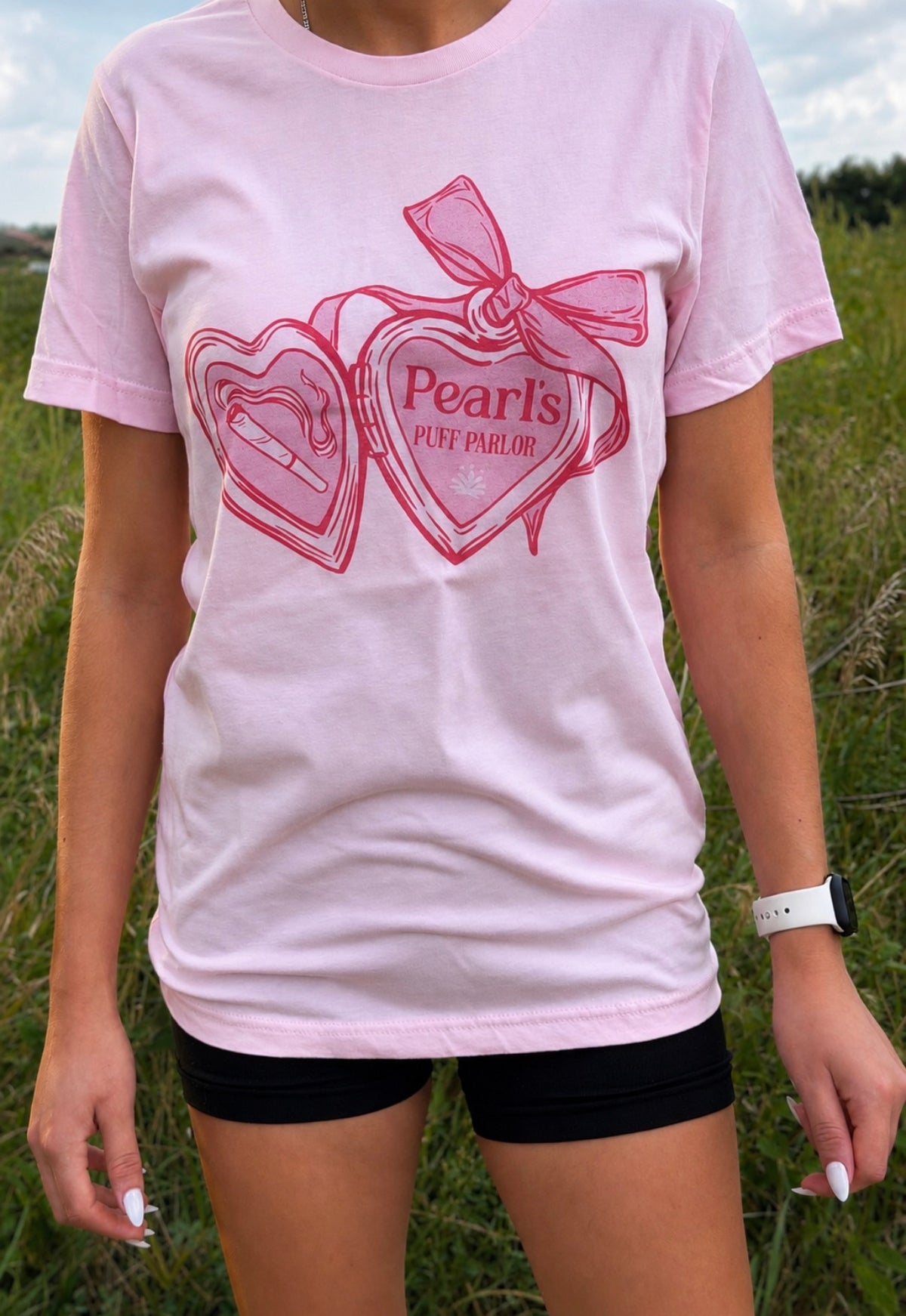 Pink t-shirt printed with a red heart-shaped locket design and 'Pearl's Puff Parlor' text, worn outdoors in a grassy field.