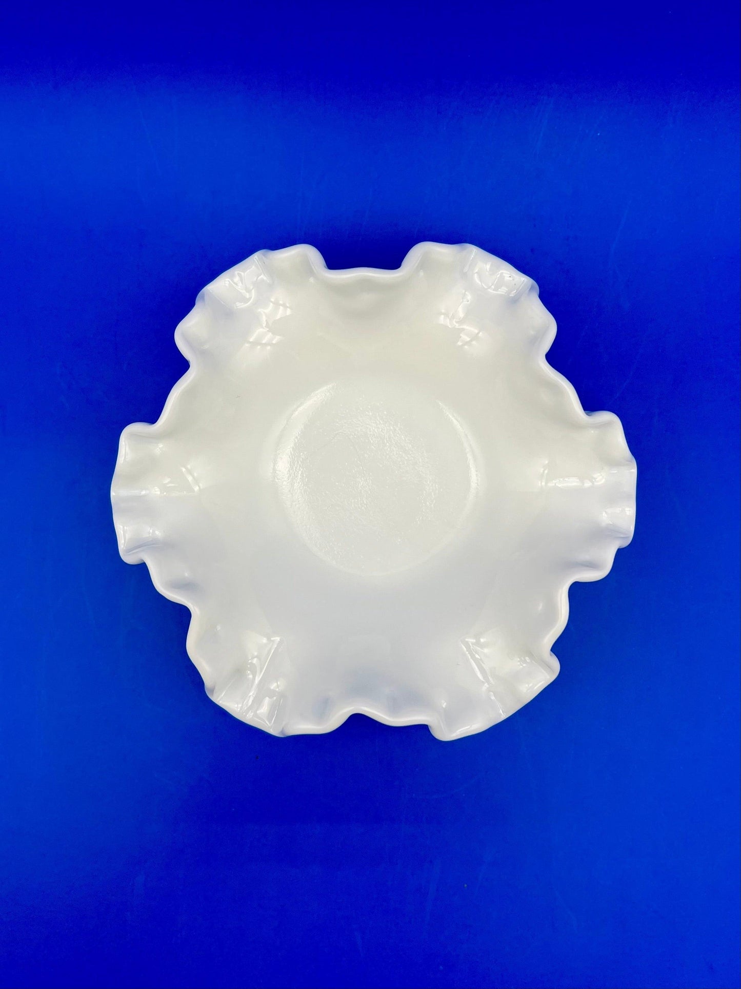 Top-down view of the same white scalloped milk glass dish showing its ruffled edge, blue background.