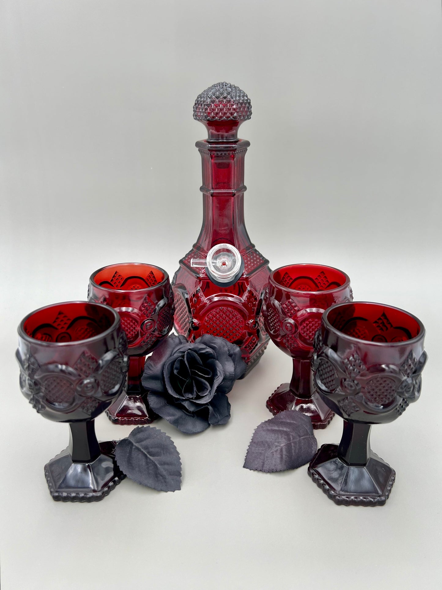 Ruby-red vintage Avon Cape Cod decanter set, transformed into a sophisticated water smoking pipe, accompanied by four matching cordial glasses, all arranged elegantly with a grey backdrop, and a subtle, black rose accent.