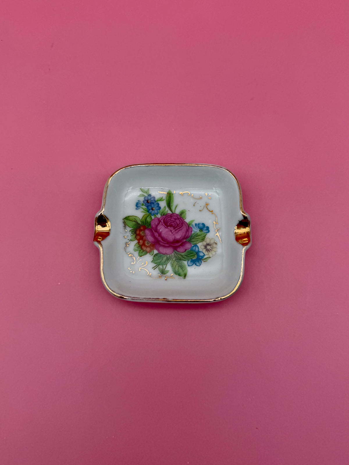Square white ashtray with gold handles, hand-painted with a pink rose and small blue flowers, pink background.