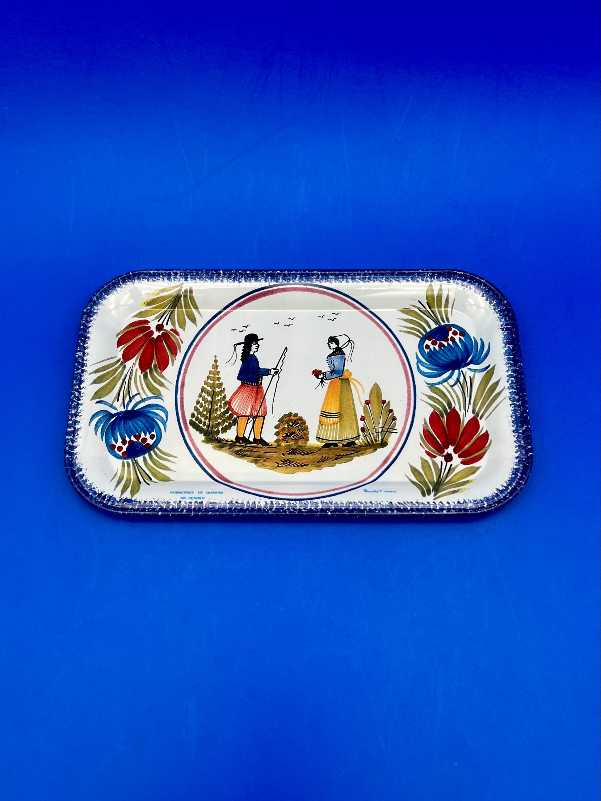 Rectangular tin tray with a Quimper-style illustration of two figures in traditional Breton dress, framed by a colorful floral border, blue background.