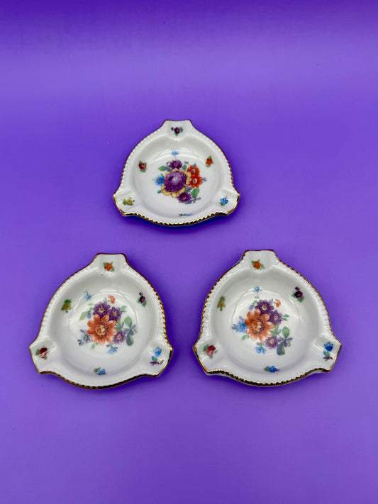 Trio of rounded triangular porcelain ashtrays with rose bouquets and gold beaded edges, purple background