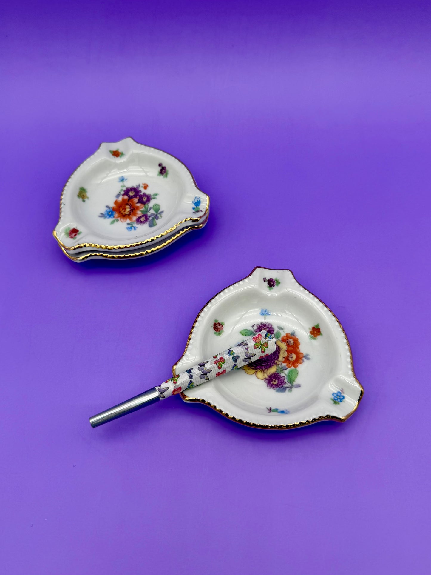 Two rounded triangular rose-patterned porcelain ashtrays with a cigarette pick resting across one, purple background