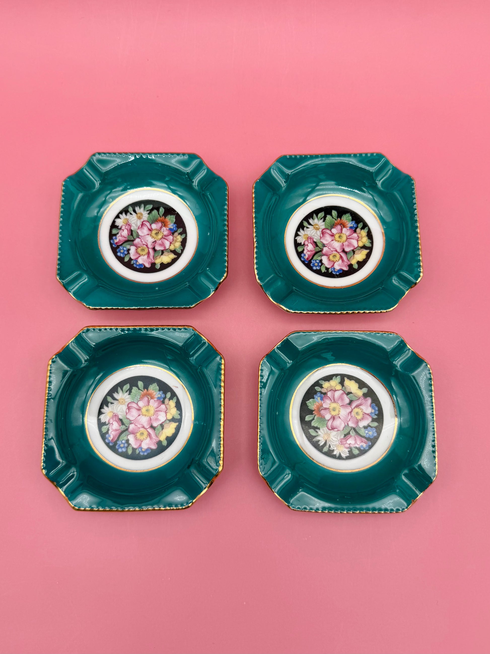 Four square teal porcelain ashtrays with colorful floral medallion centers arranged in a grid, pink background