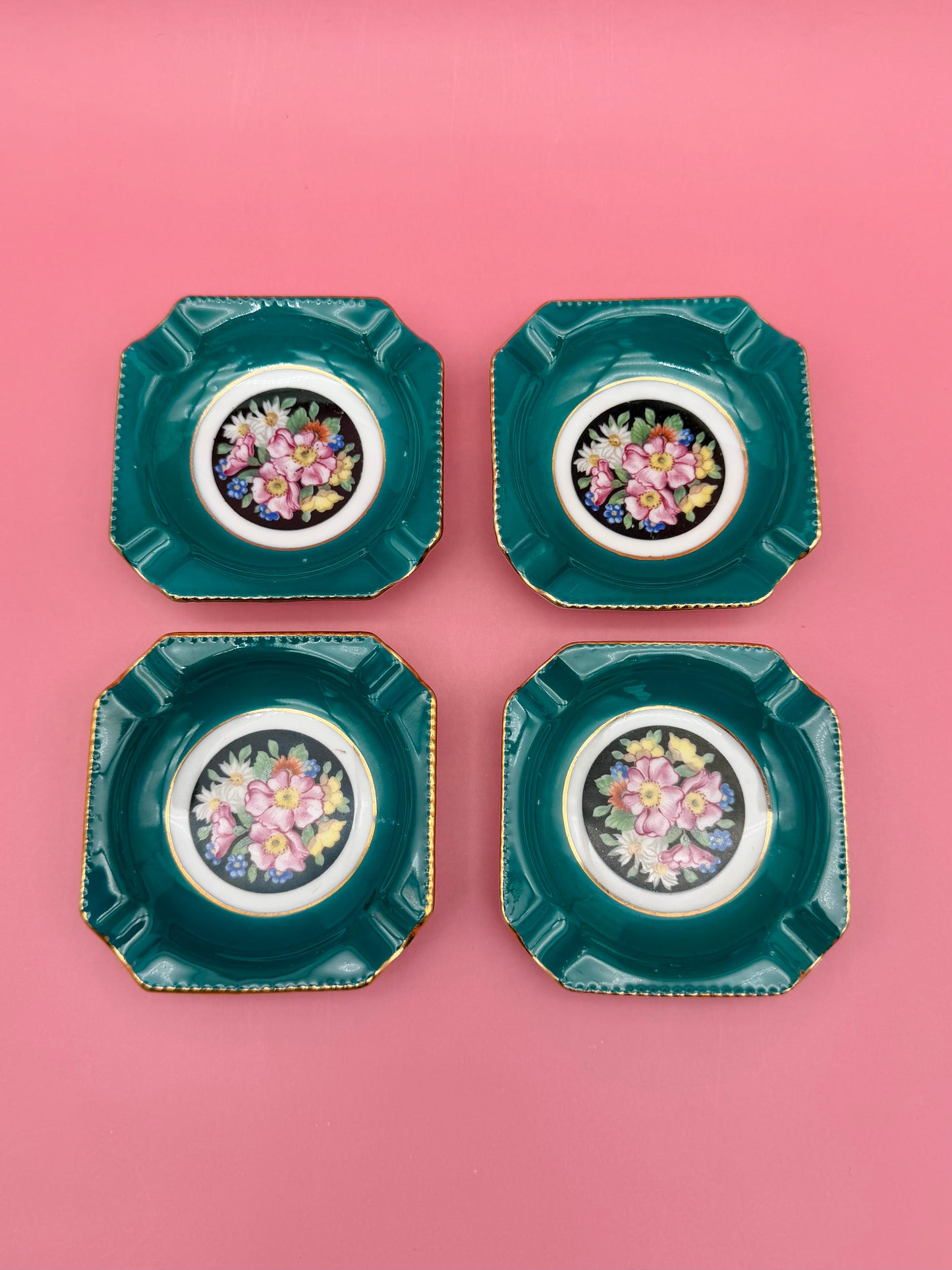 Four square teal porcelain ashtrays with colorful floral medallion centers arranged in a grid, pink background