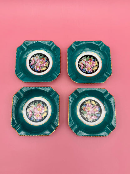Four square teal porcelain ashtrays with colorful floral medallion centers arranged in a grid, pink background
