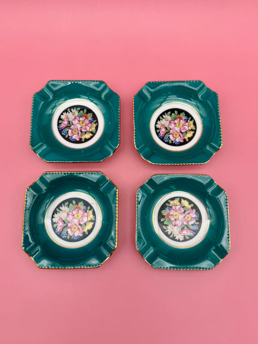 Four square teal porcelain ashtrays with colorful floral medallion centers arranged in a grid, pink background