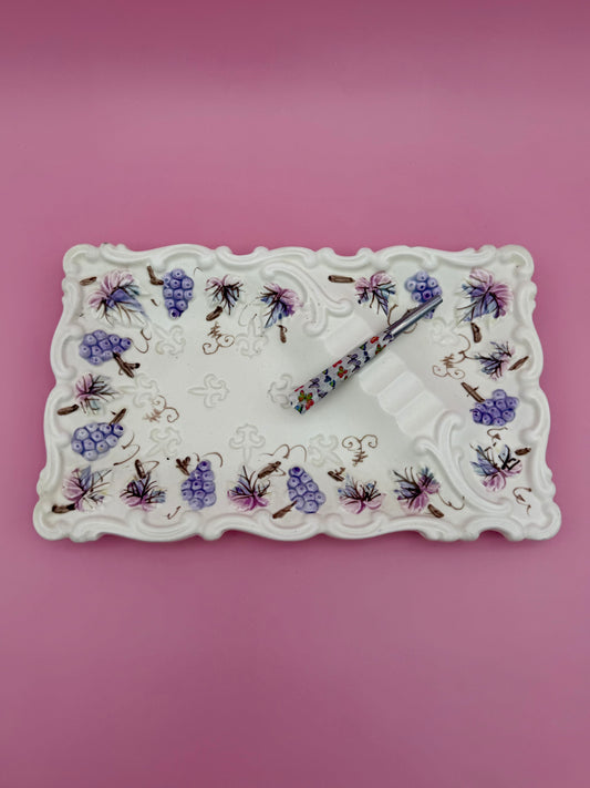 Rectangular white porcelain tray with a scalloped edge and a hand-painted purple grape and floral vine pattern, glass cigarette pick resting across it, pink background.