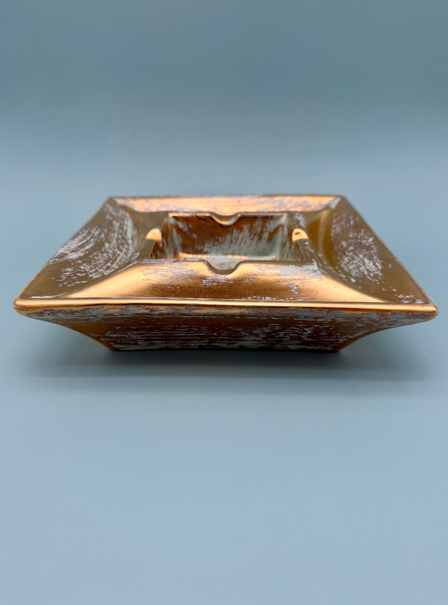 Square gold and copper pottery ashtray shown from a side profile, gray background