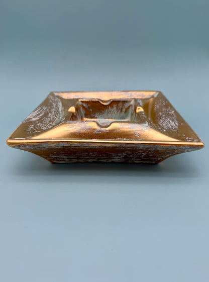 Square gold and copper pottery ashtray shown from a side profile, gray background