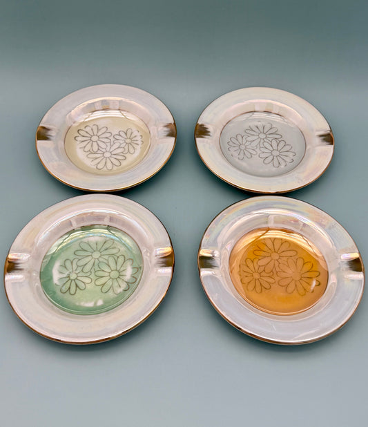 Four round glass ashtrays in different tinted colors (clear, gray, green, and amber), each with an embossed daisy pattern in the center and a gold rim, gray background.