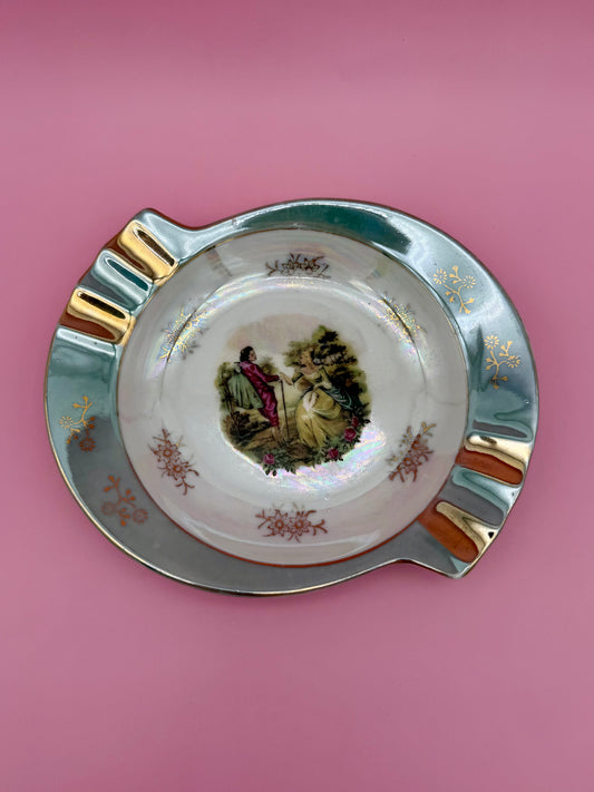 Scalloped iridescent lustreware ashtray with a painted Victorian courting-couple scene in the center and gold trim, pink background.