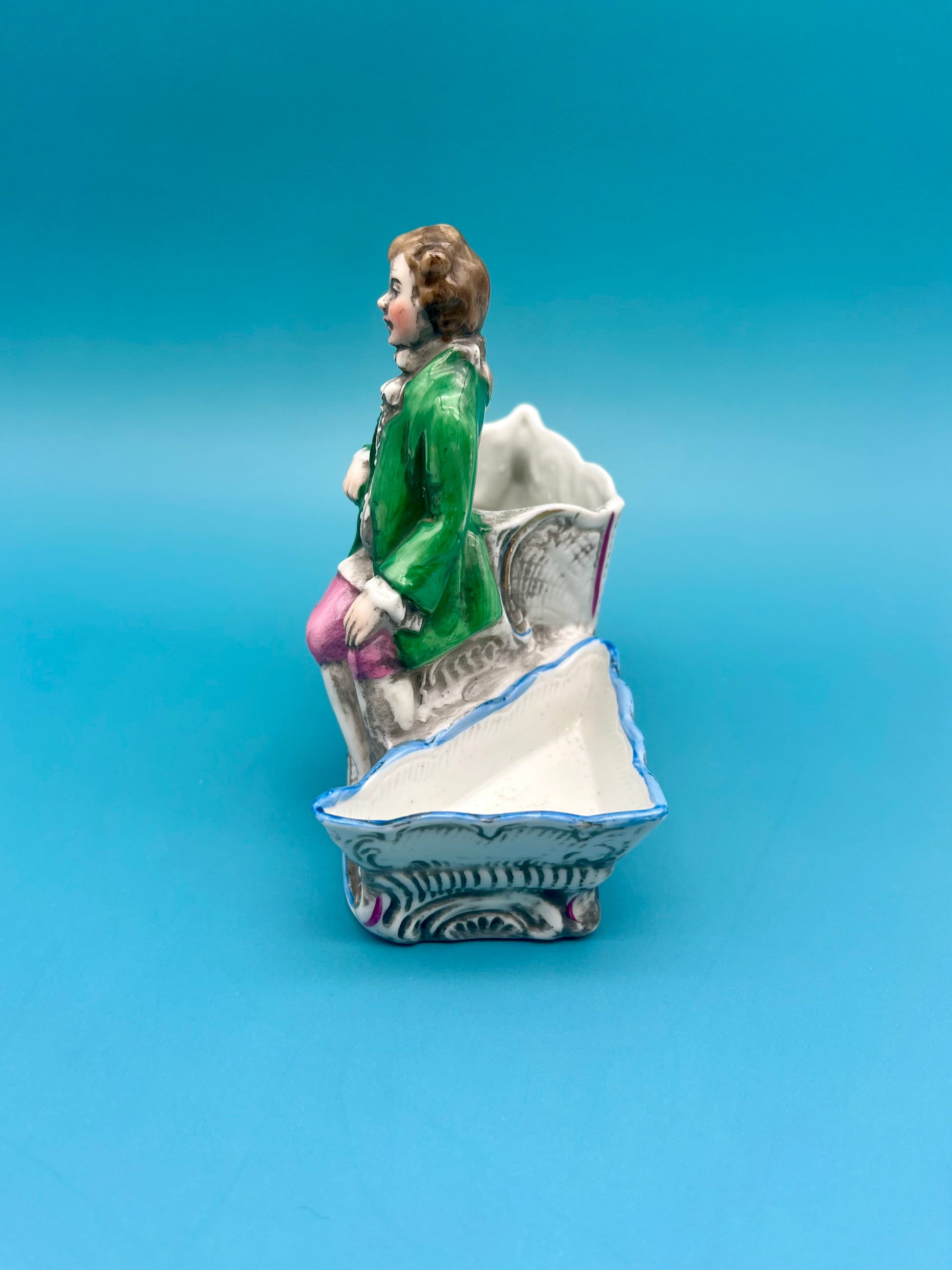 Side profile view of the Ludwig Set gentleman figurine seated on the scrollwork base, teal background.