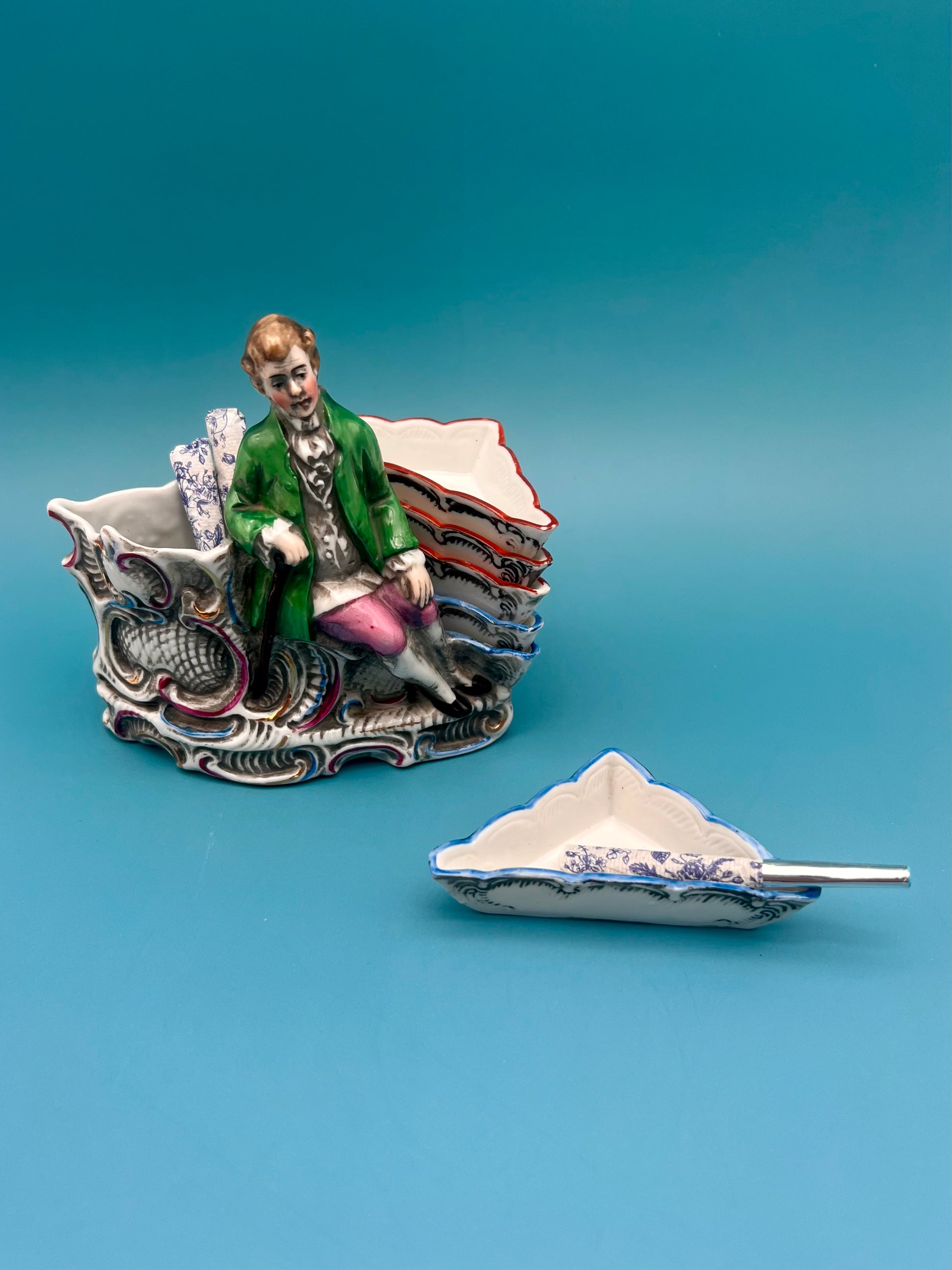 Ludwig Set porcelain figurine of a gentleman in an 18th-century green coat seated on an ornate rococo scrollwork base, positioned beside a matching ashtray, teal background.
