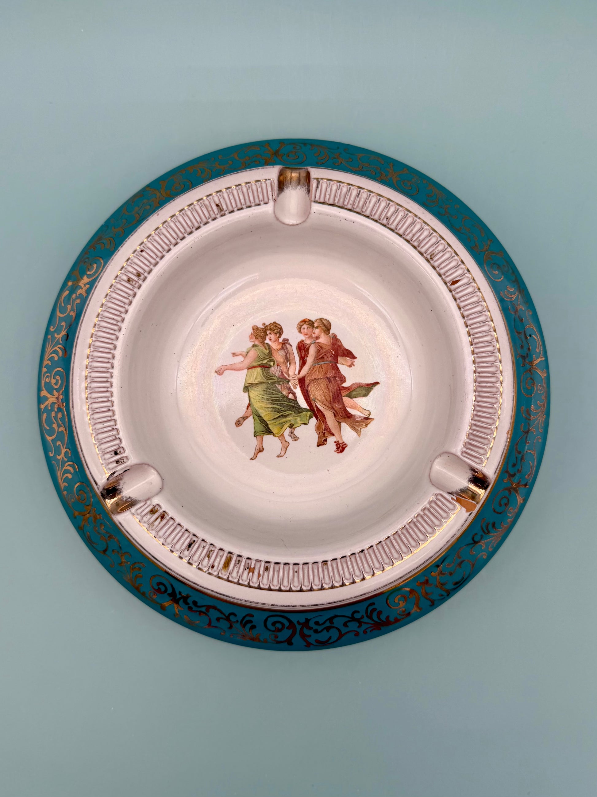 Round turquoise porcelain ashtray with a dancing figures medallion and ornate gold scrollwork, gray background