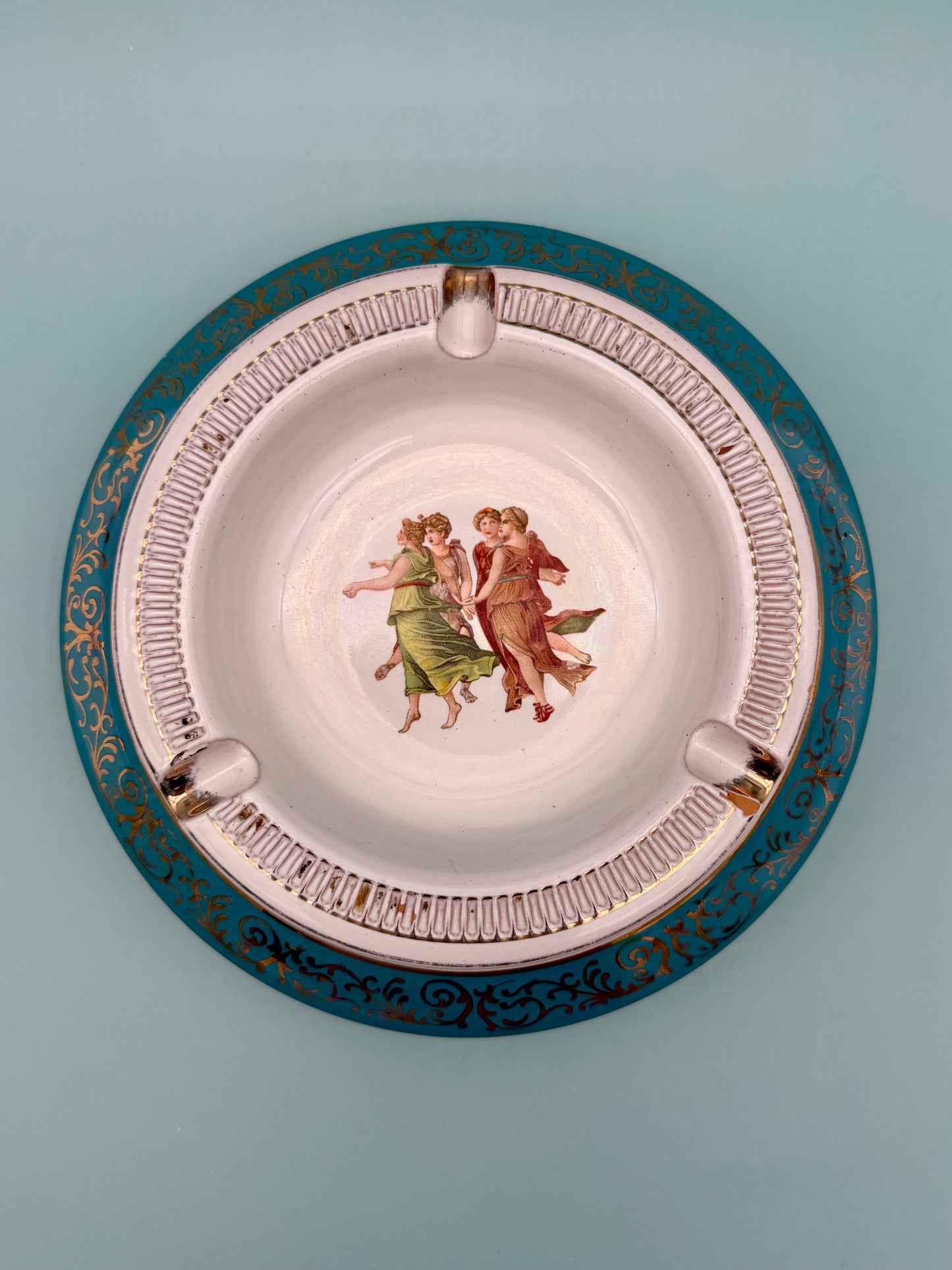 Round turquoise porcelain ashtray with a dancing figures medallion and ornate gold scrollwork, gray background