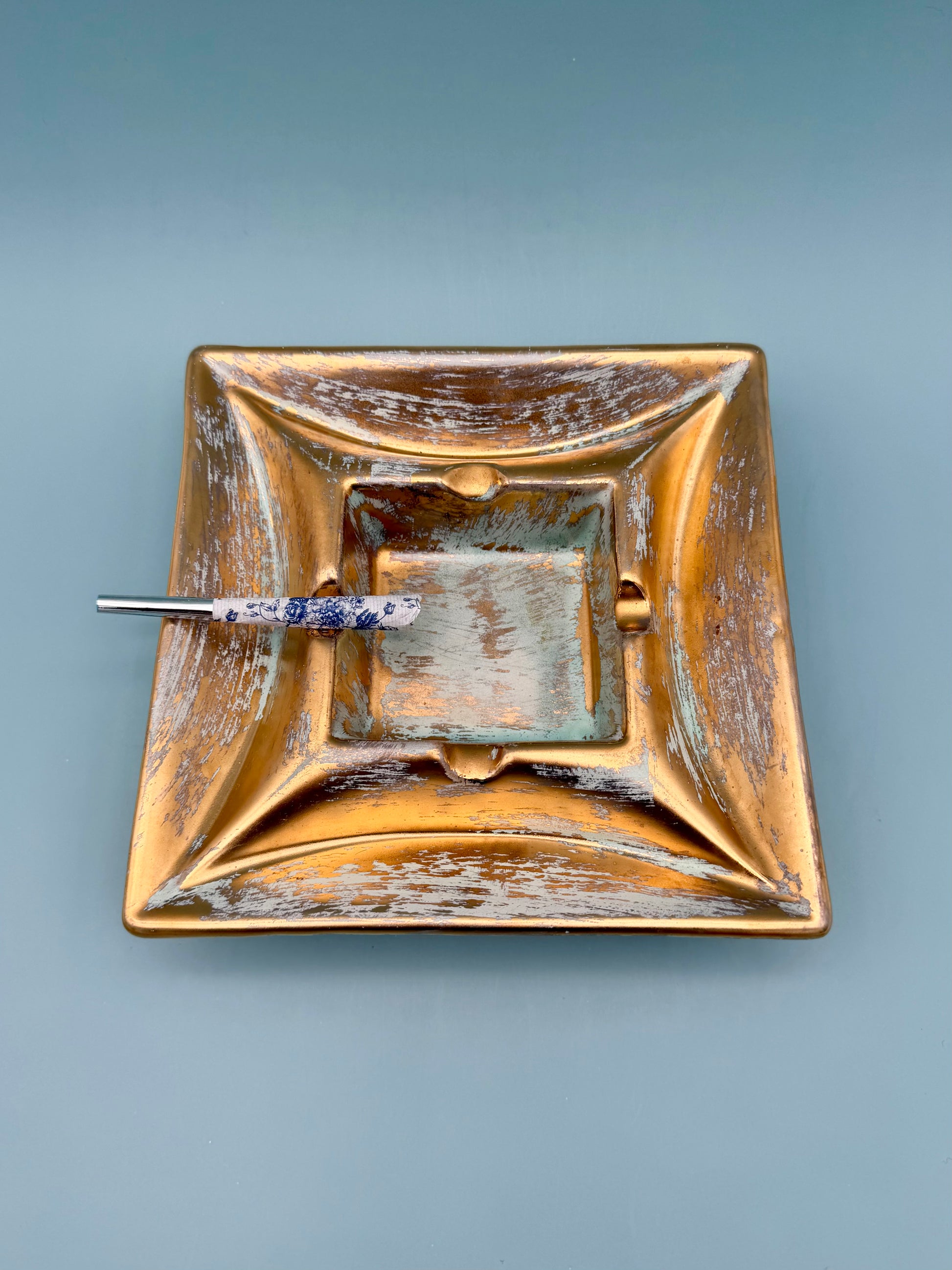 Square gold and copper toned pottery ashtray with blue patina accents and a cigarette pick resting inside, gray background
