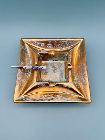 Square gold and copper toned pottery ashtray with blue patina accents and a cigarette pick resting inside, gray background