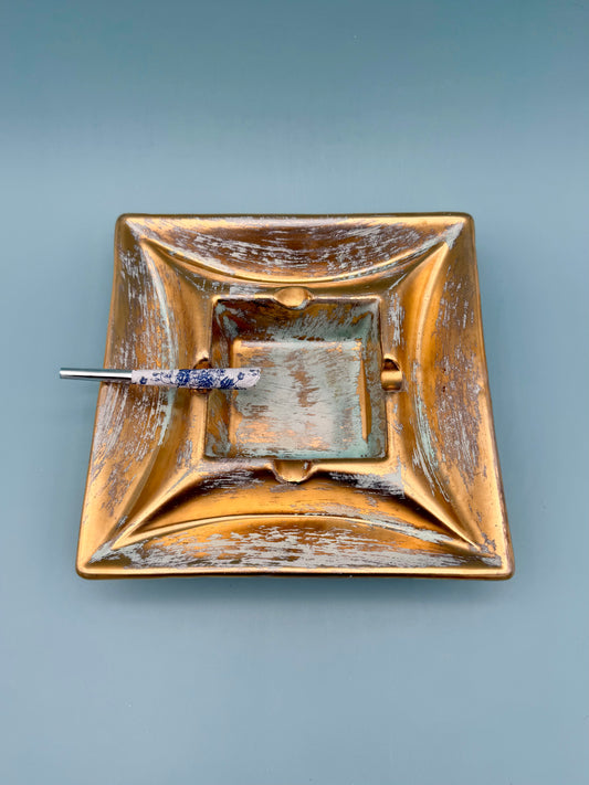 Square gold and copper toned pottery ashtray with blue patina accents and a cigarette pick resting inside, gray background