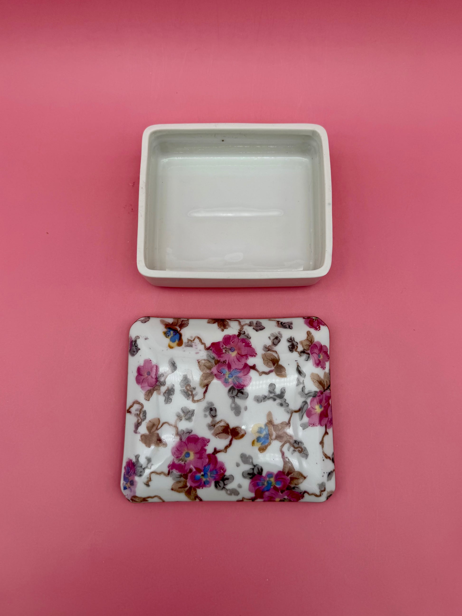 Open empty porcelain stash box beside its floral-painted lid, pink background