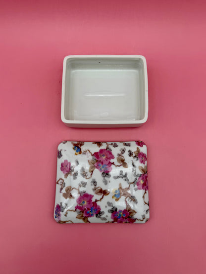 Open empty porcelain stash box beside its floral-painted lid, pink background