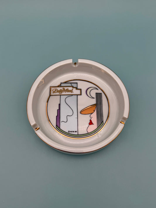 Round white ceramic ashtray with a printed illustration of a stylized bedroom vanity scene (dresser, chair, and standing lamp) in the center, gold rim, viewed from above, teal-blue background.