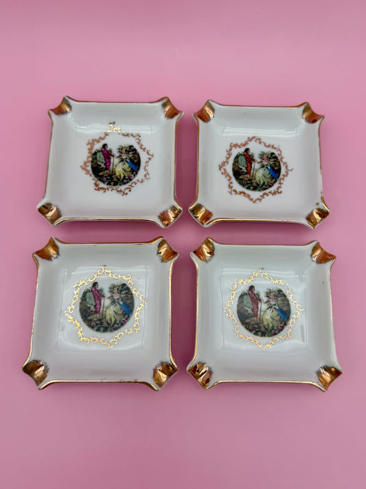 Four square white ceramic ashtrays with gold corner accents, each painted with an oval Victorian courting-couple medallion, arranged in a 2x2 grid, pink background.
