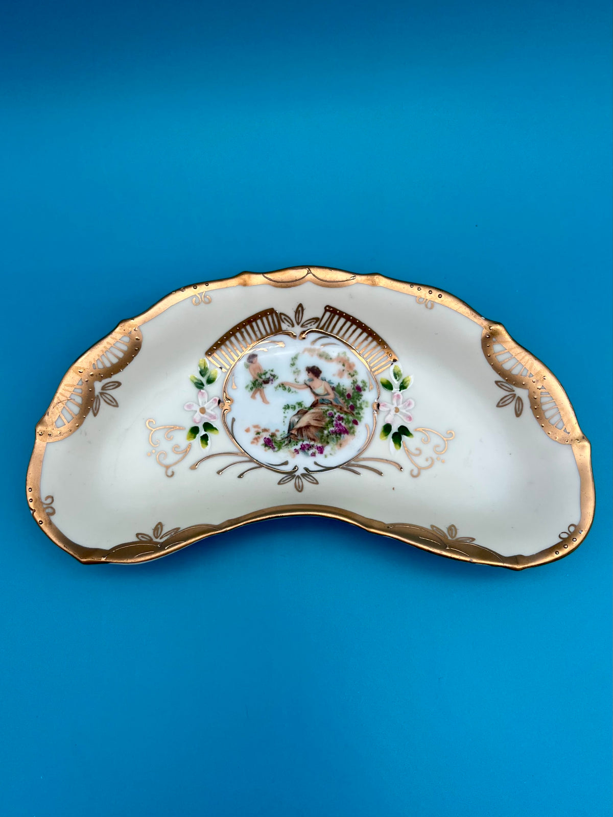 Close-up of the Arabella ashtray showing the gold-trimmed crescent shape and painted center medallion, blue background.