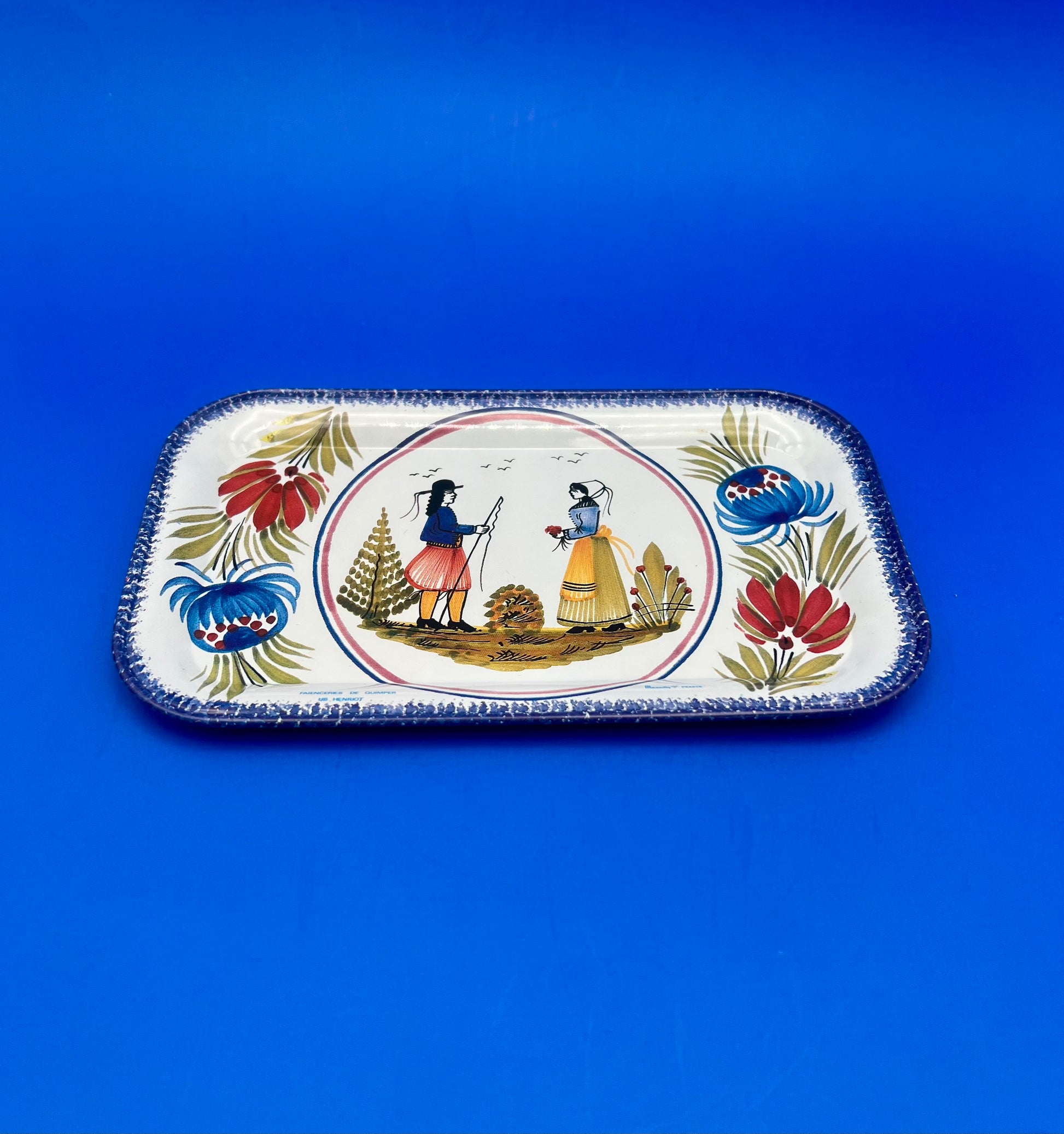 Angled view of the Camille Tray showing the Quimper-style folk illustration and floral border, blue background.