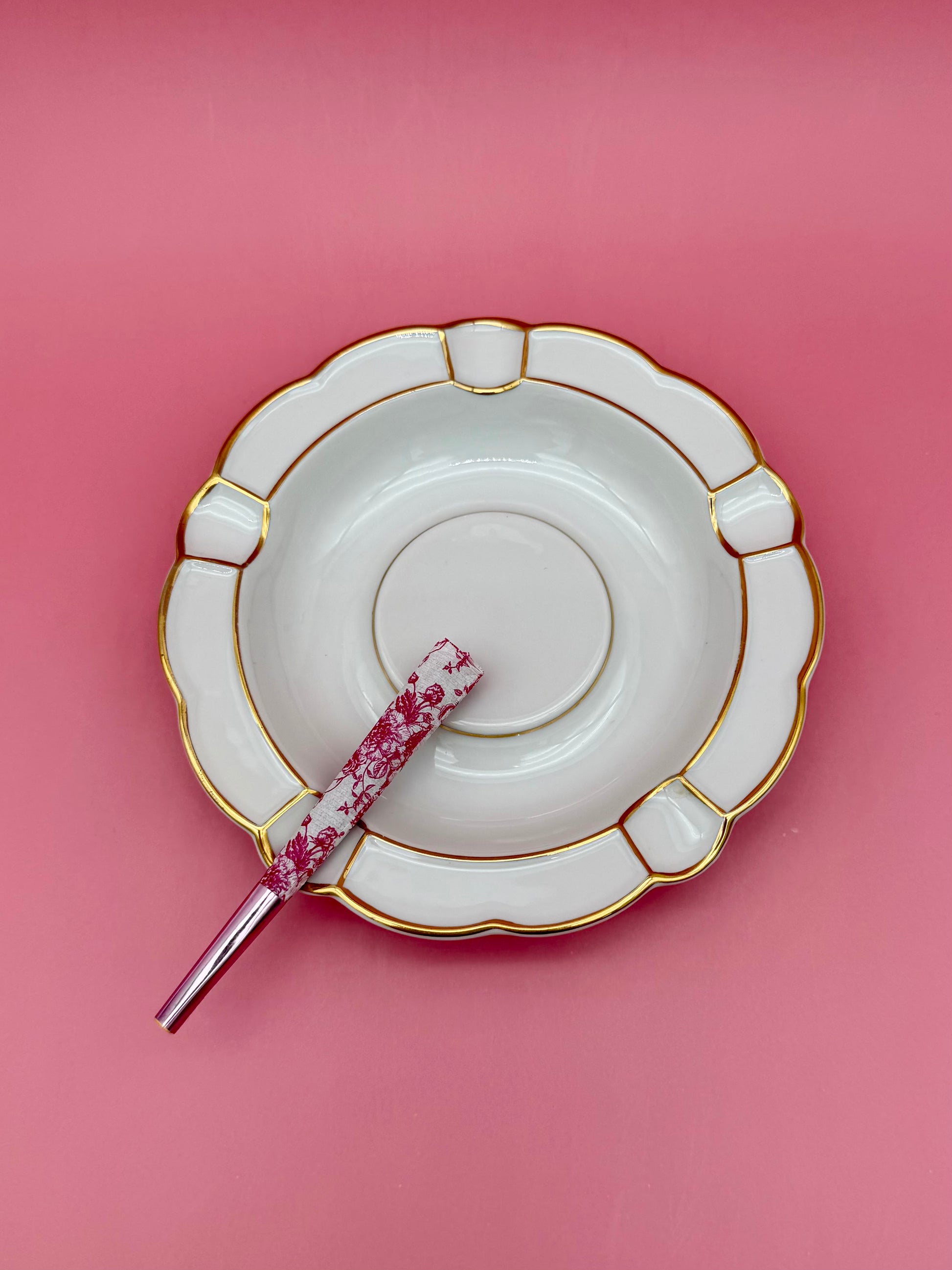 Round white porcelain ashtray with a paneled scalloped border and gold detailing, glass cigarette pick resting across it, pink background