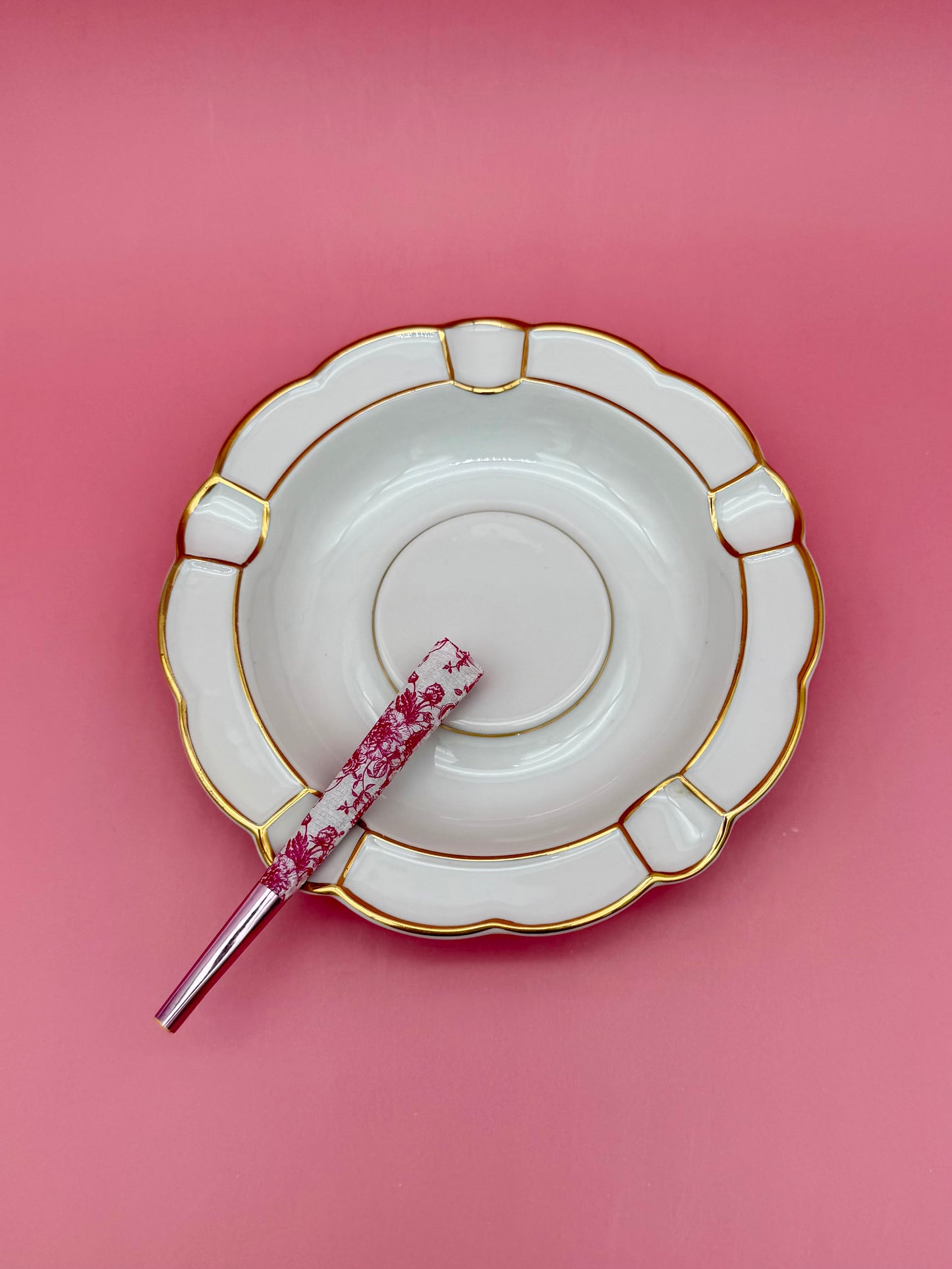 Round white porcelain ashtray with a paneled scalloped border and gold detailing, glass cigarette pick resting across it, pink background