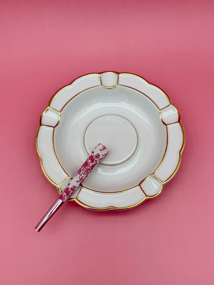 Round white porcelain ashtray with a paneled scalloped border and gold detailing, glass cigarette pick resting across it, pink background