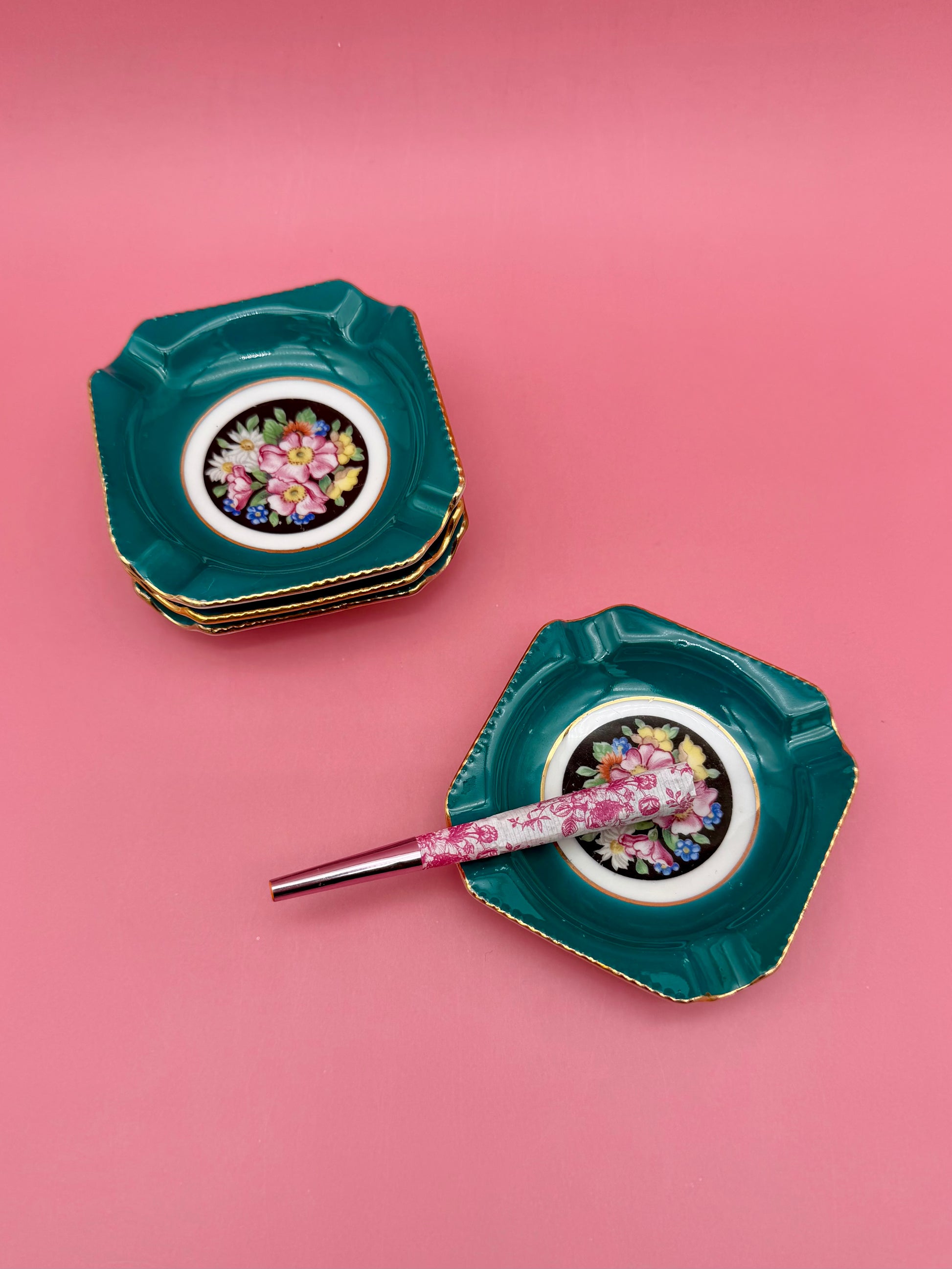 Stacked teal floral medallion ashtrays with one tipped forward and a cigarette pick resting beside it, pink background