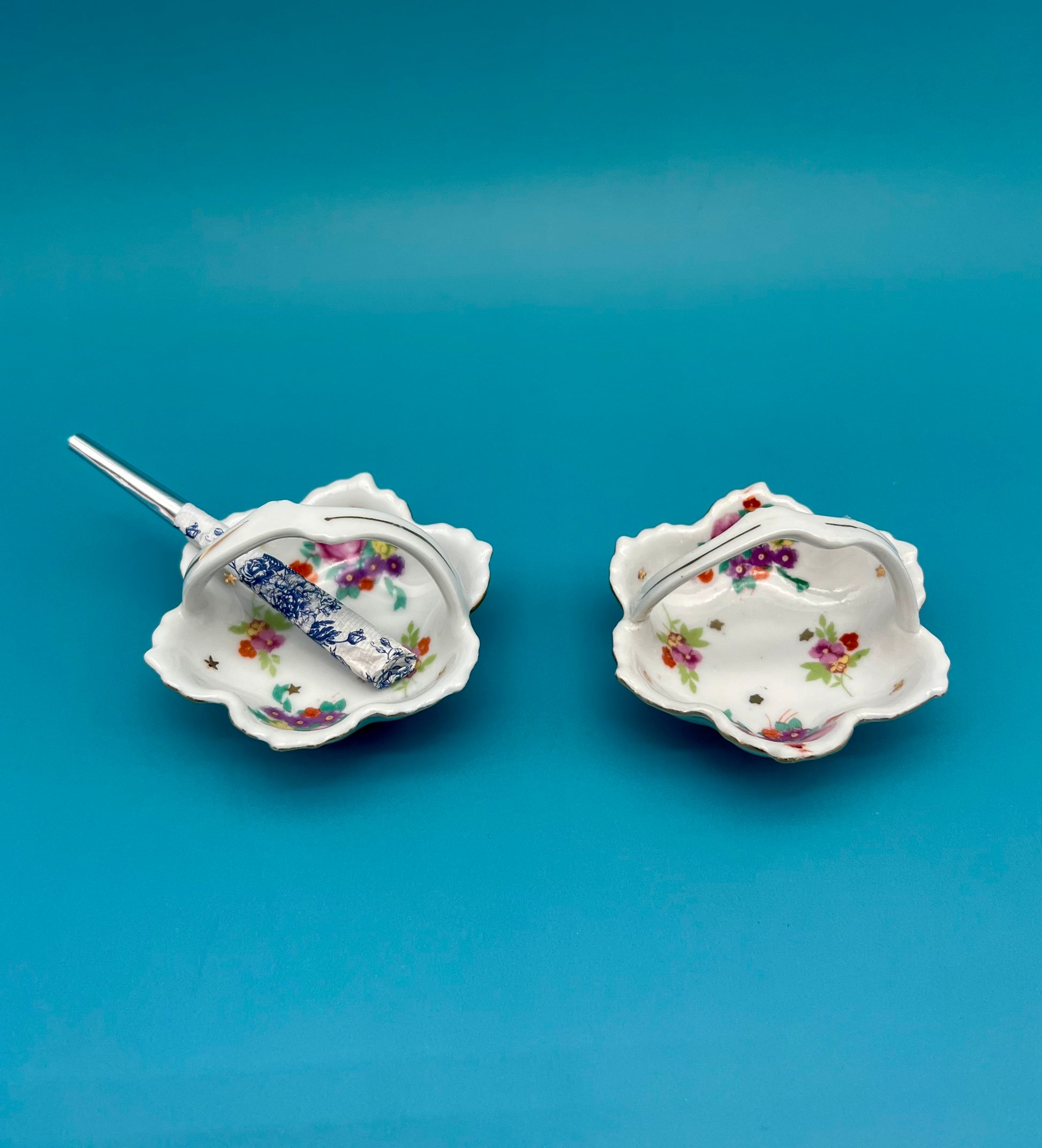 Two small scalloped porcelain ashtrays from the Reagan Set with colorful floral sprays and gold star accents, glass cigarette pick resting in one, teal background.