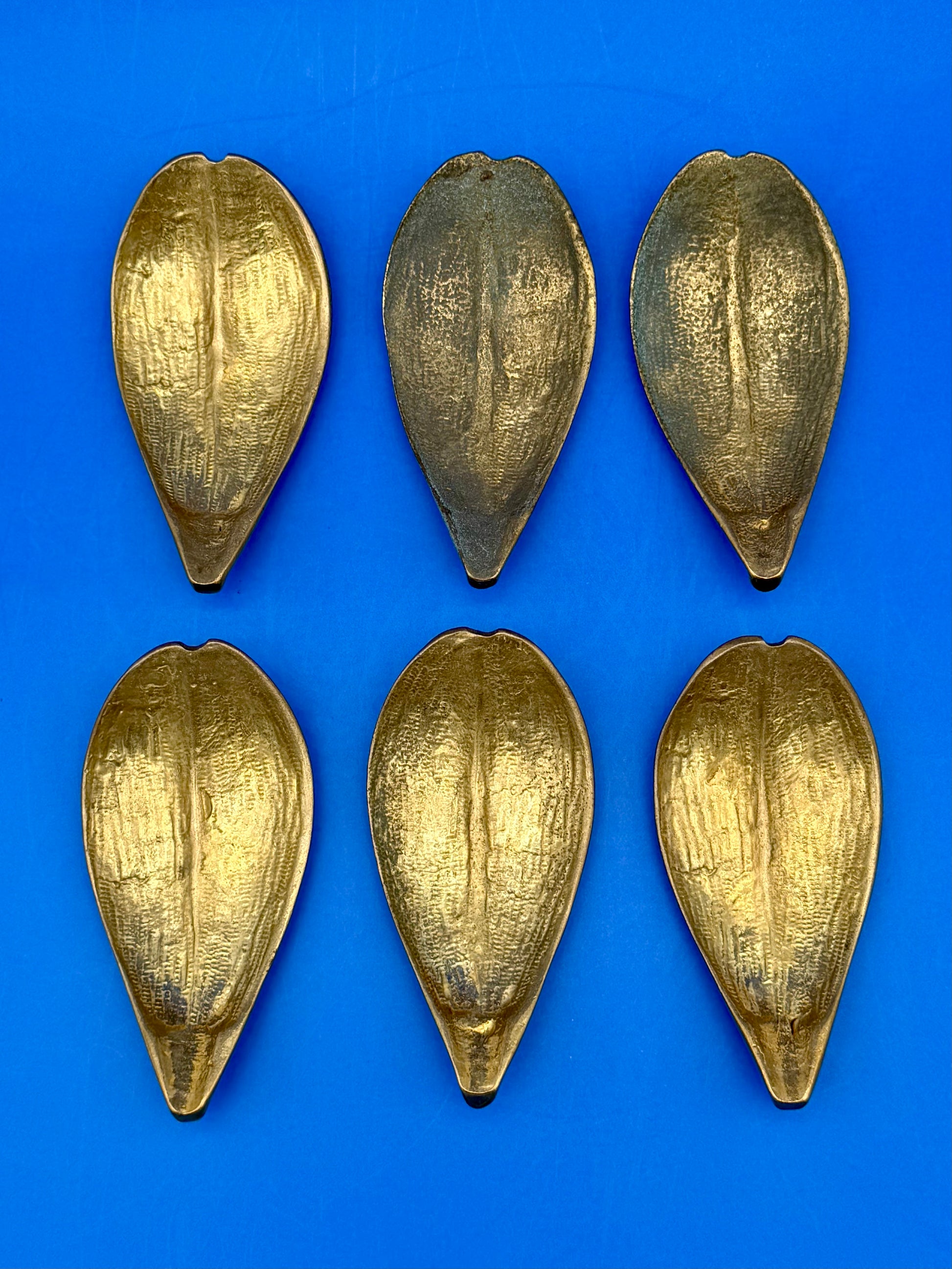 Six brass leaf-shaped petal ashtrays with textured detailing arranged in two rows, shown from a different angle, blue background