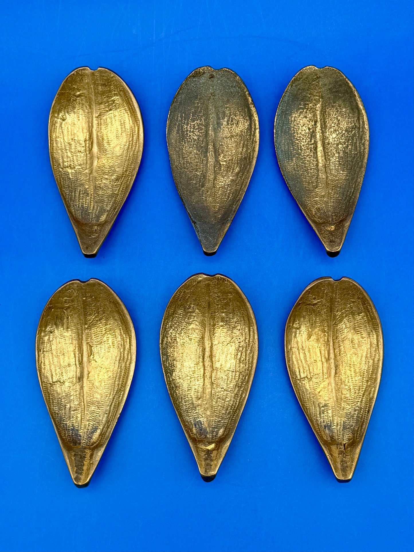 Six brass leaf-shaped petal ashtrays with textured detailing arranged in two rows, shown from a different angle, blue background