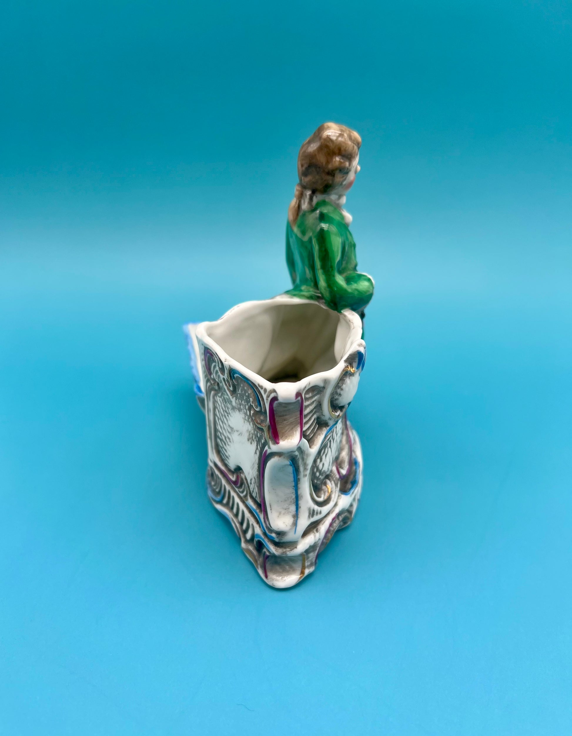 Back view of the Ludwig Set figurine showing the seated gentleman from behind within the ornate scrollwork holder, teal background.
