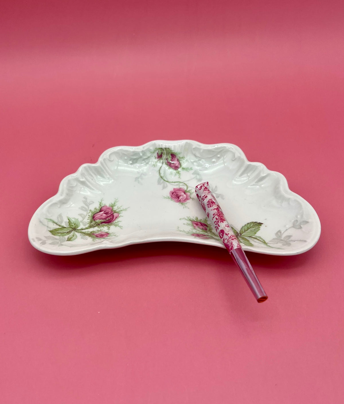Crescent-shaped white bone china ashtray with a delicate pastel pink floral pattern, glass cigarette pick resting across it, pink background.