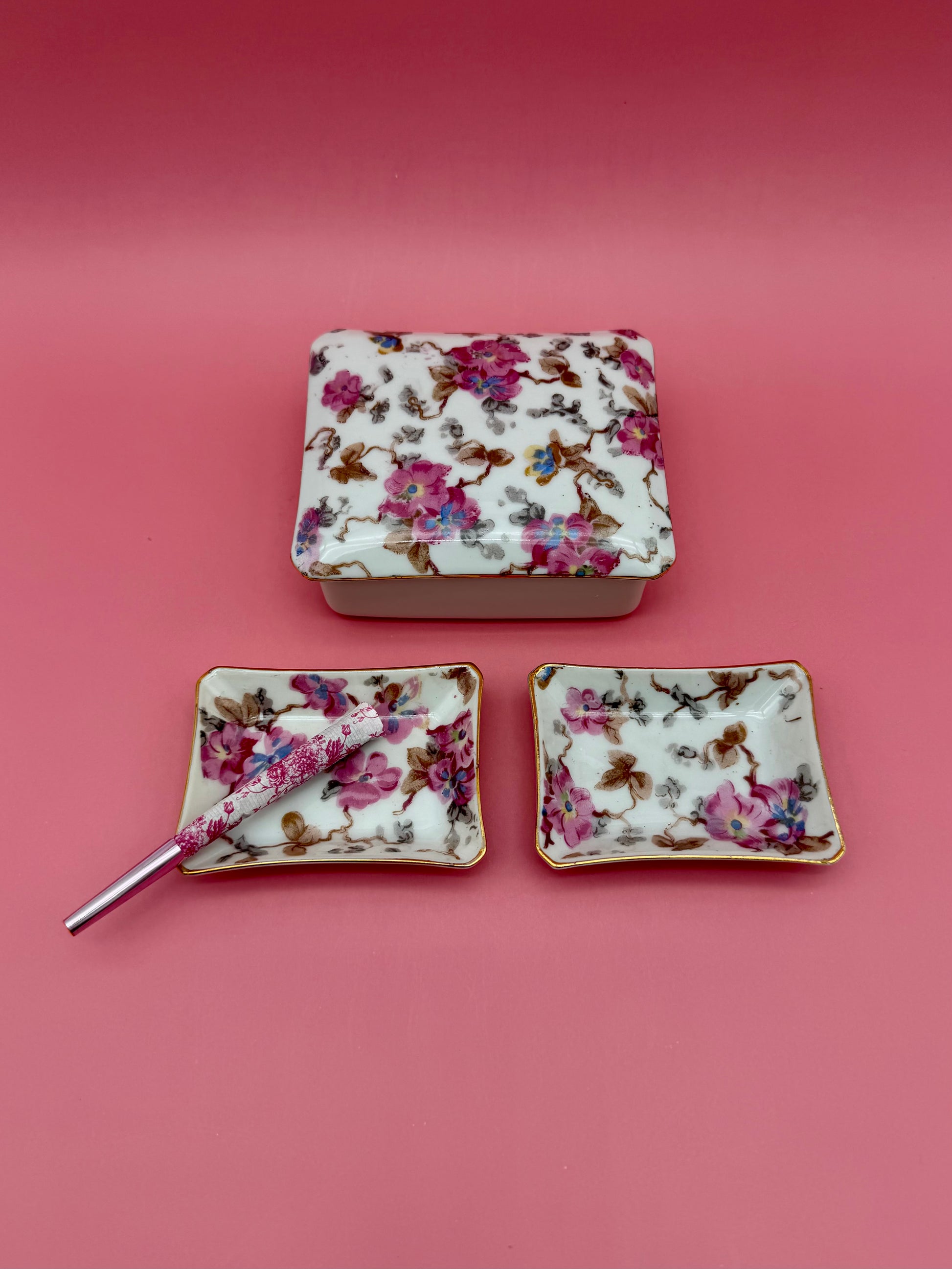 Rectangular floral porcelain stash box beside two matching floral ashtrays with a cigarette pick, dark pink background