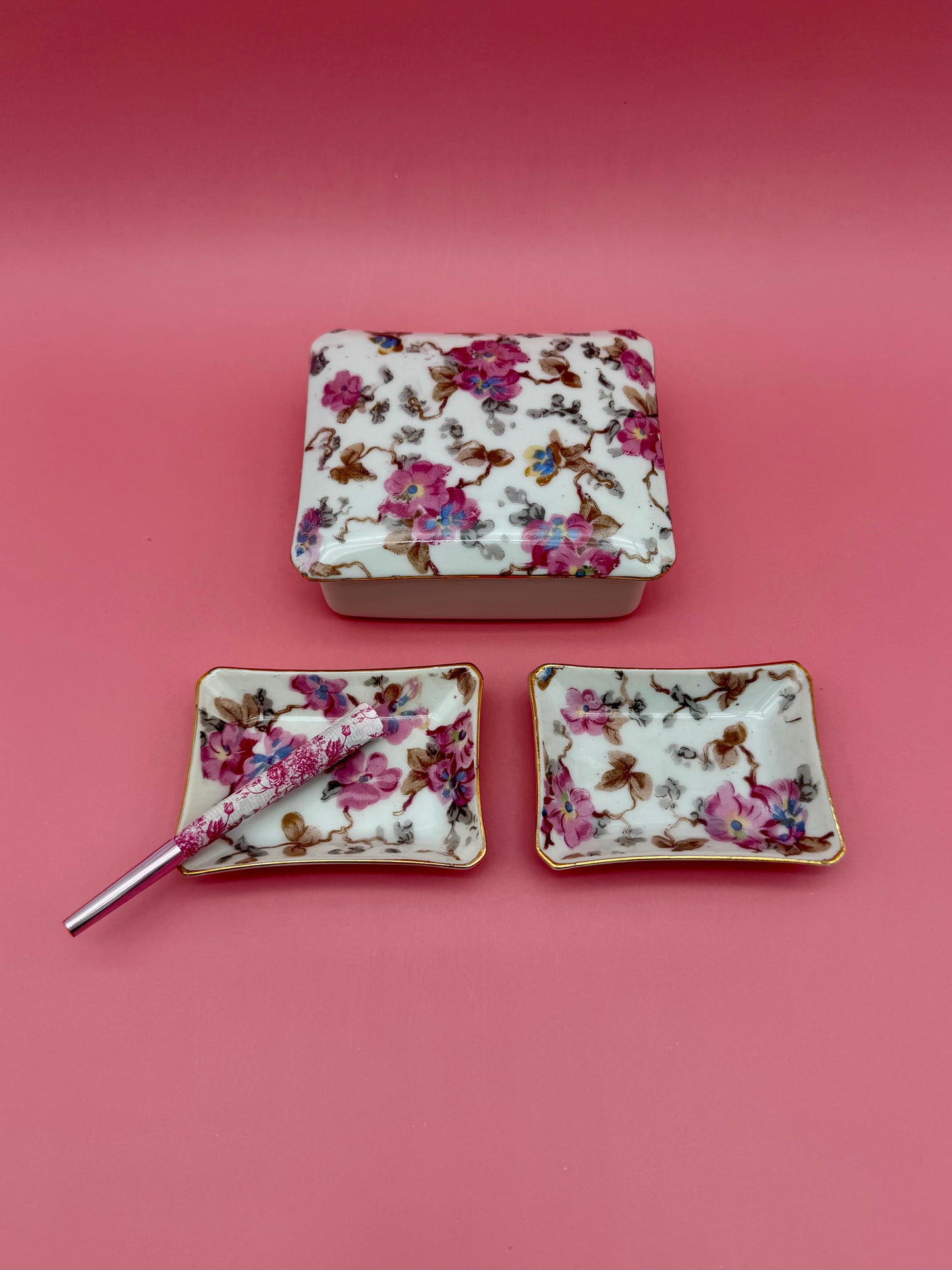 Rectangular floral porcelain stash box beside two matching floral ashtrays with a cigarette pick, dark pink background