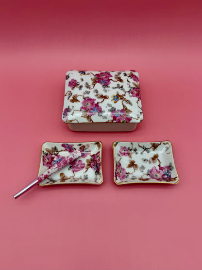 Rectangular floral porcelain stash box beside two matching floral ashtrays with a cigarette pick, dark pink background