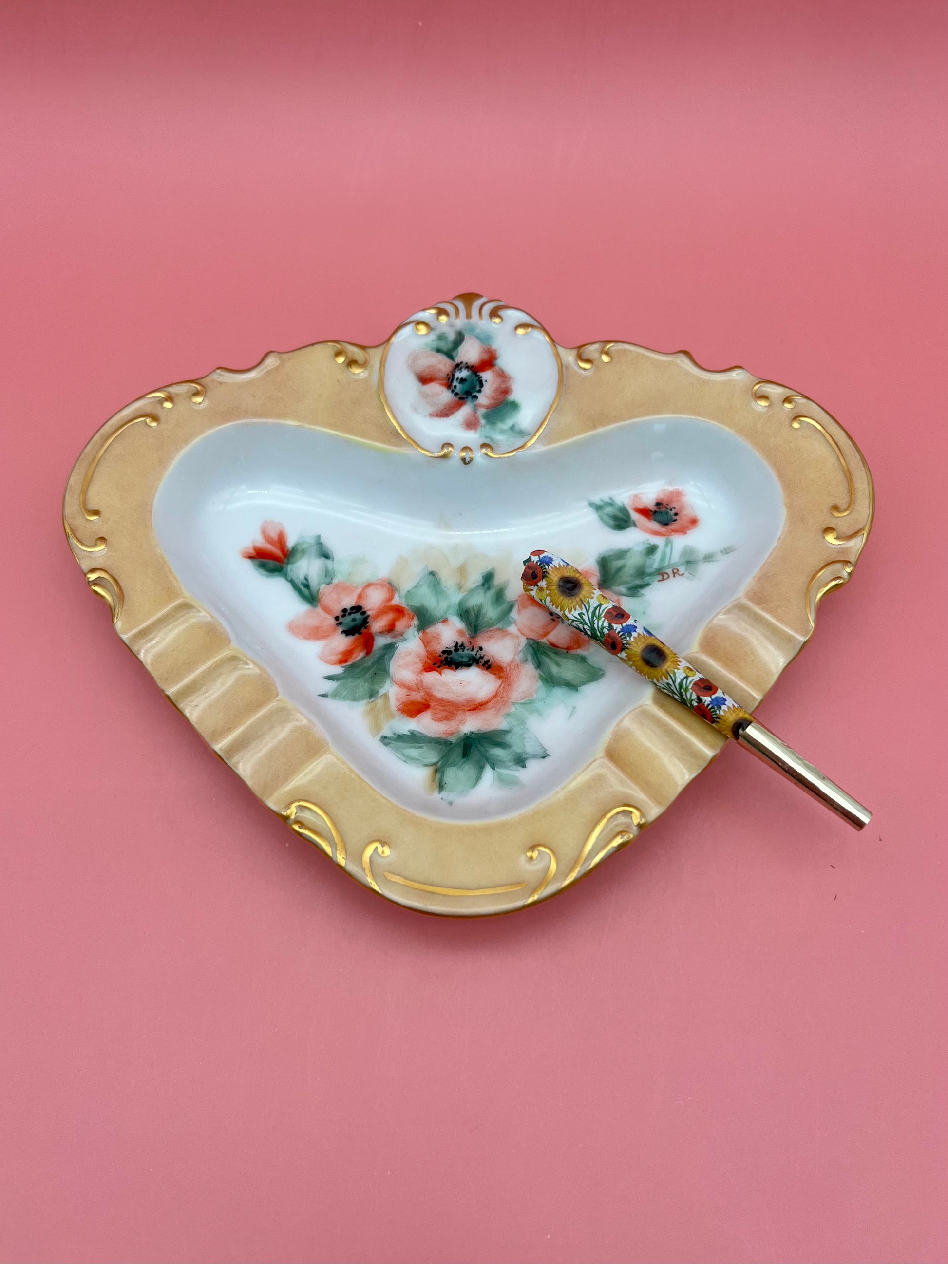 Front view of a triangular porcelain ashtray with a gold scrollwork border and hand-painted coral pink florals, glass cigarette pick resting across it, pink background.