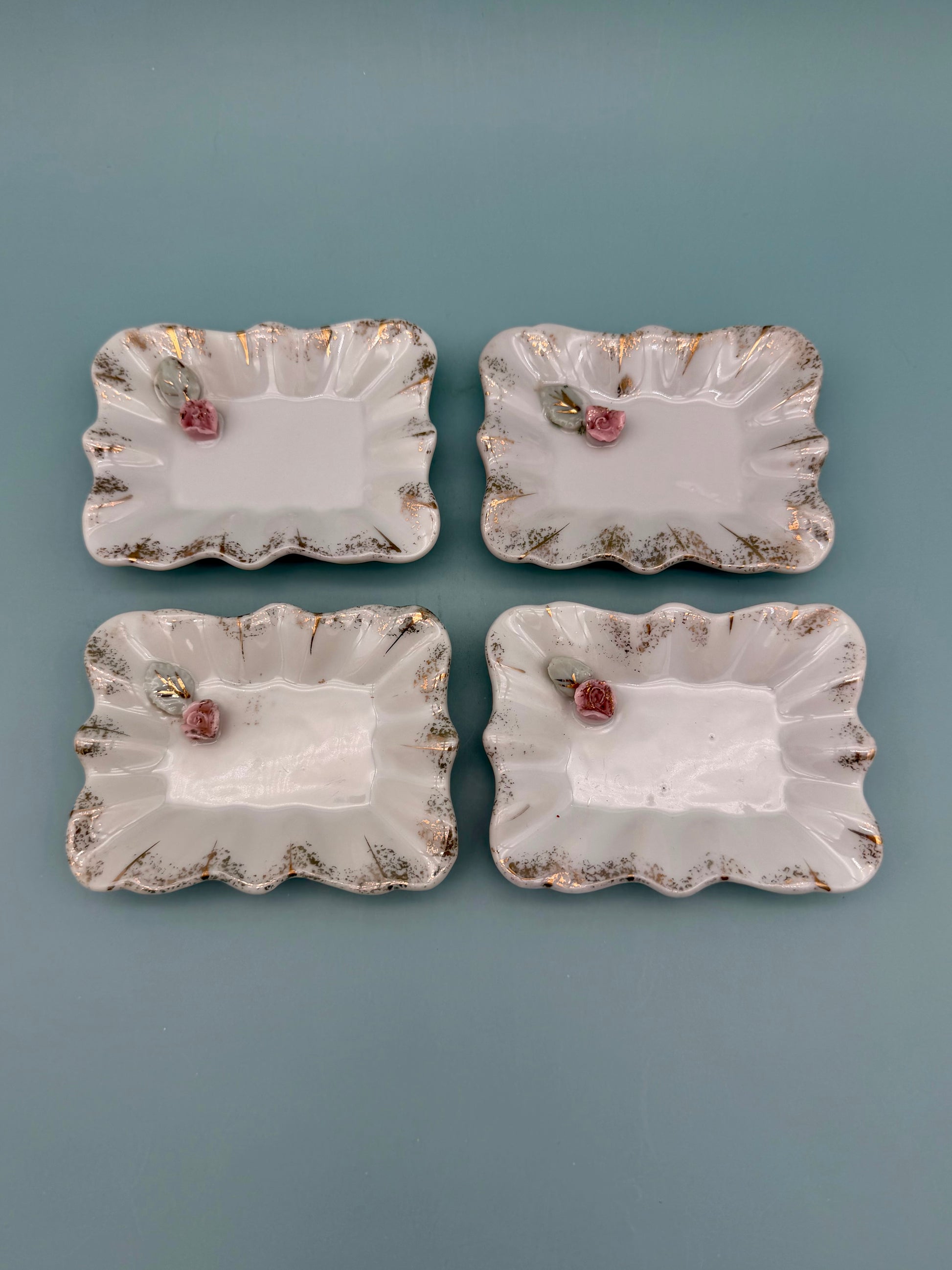 Four scalloped porcelain mini trays with pink roses and gold accents arranged in a square, teal background