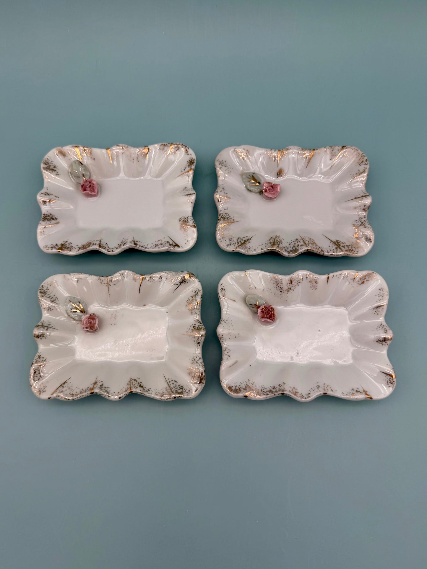 Four scalloped porcelain mini trays with pink roses and gold accents arranged in a square, teal background