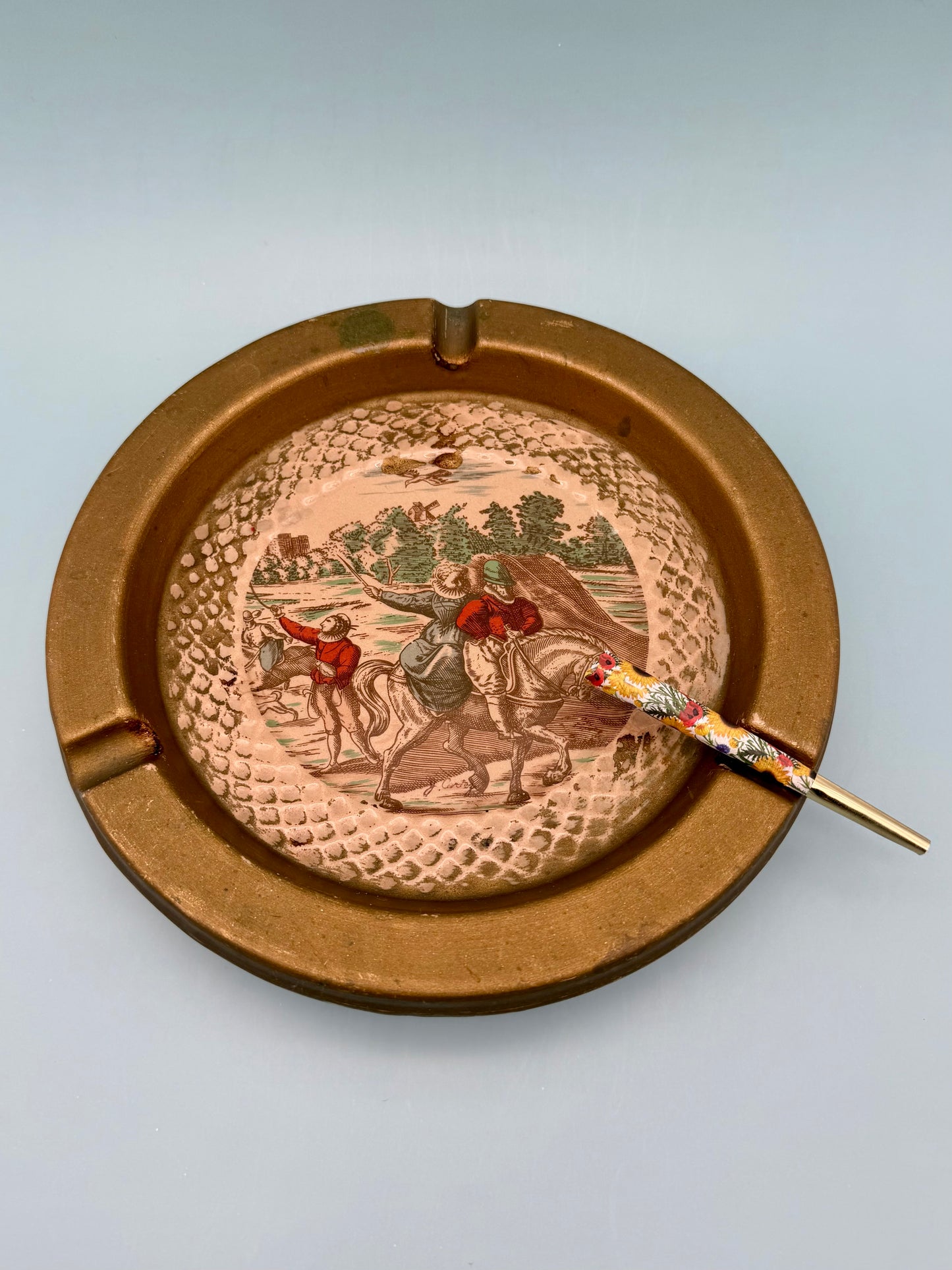 Round Florentine ashtray with hand-painted horseback rider landscape scene and gold toned frame, with a cigarette pick, gray background