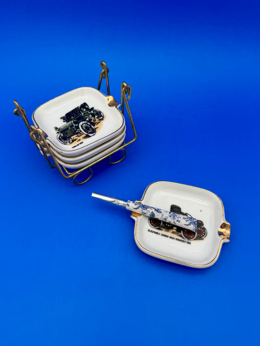 Stack of square white ceramic ashtrays with gold corner handles, each painted with a different vintage automobile illustration, seated in a gold wire caddy, blue cigarette pick resting nearby, blue background.
