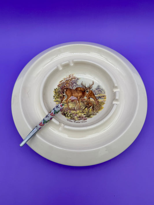 Round white ceramic ashtray with a wide rim and a printed deer scene (buck and doe in a woodland setting) at the center, glass cigarette pick resting across it, purple background.