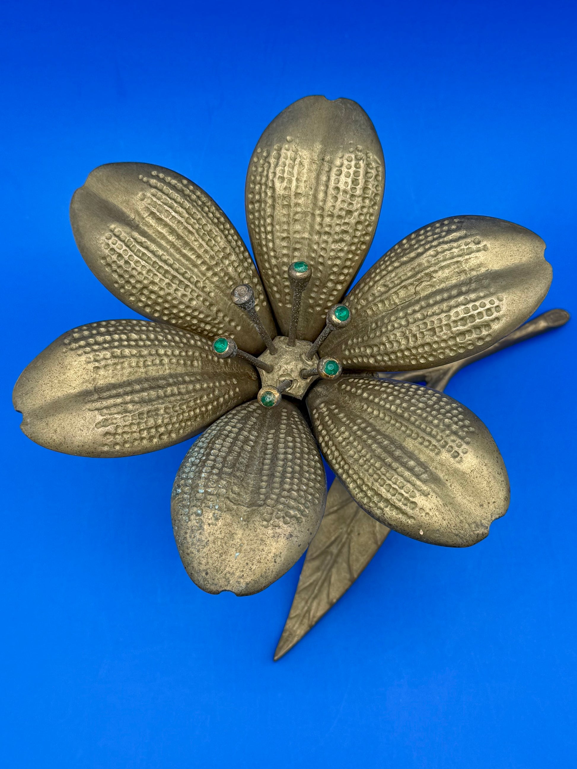 Top-down view of the brass flower ashtray showing all petals and green jewel centers, blue background