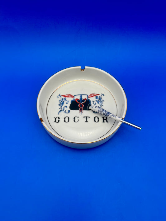 Round white porcelain ashtray with gold trim and a doctor's bag and caduceus emblem reading 'DOCTOR,' glass cigarette pick resting across it, blue background.
